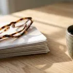 Reading glasses resting on a stack of papers beside a cup of black coffee on a sunlit desk
